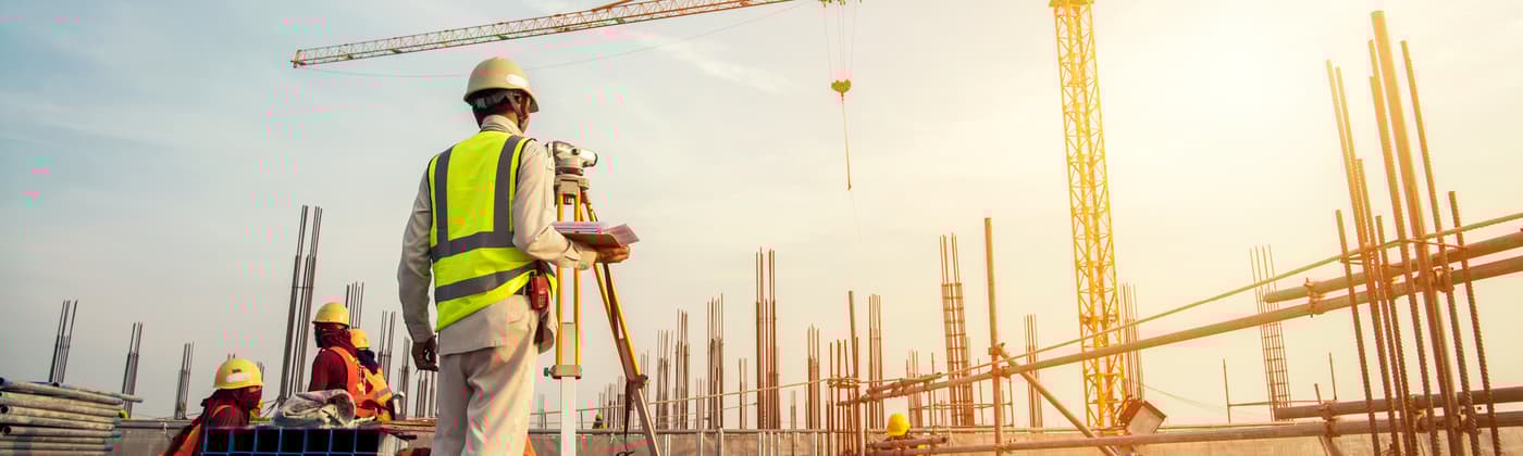 Construction site worker with hard hat and surveying equipment looking up at crane and preparing for building materials inspection