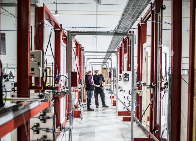 Two building products testing engineers in hard hats speaking to each other while standing centered within a series of metal testing frames in their laboratory